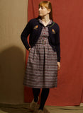 A person wears a Palava Vera Navy Robins Cardigan in navy blue organic cotton with bird embroidery over a plaid button-up dress.