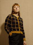 A person wears a BUTIK KUBIK Palava Vera Navy Mustard Check Cardigan, an eco-friendly navy blue button-down jacquard knit with a mustard yellow and white windowpane check pattern and solid mustard yellow trim.