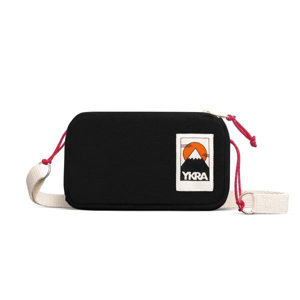 The BUTIK KUBIK YKRA travel Black is a black rectangular crossbody bag with a cream adjustable strap and red cord accents. Ideal for travel essentials, it sports a mountain patch, YKRA at the top right corner, and a durable YKK zipper for secure storage.