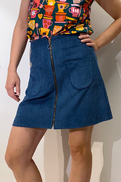 A person is wearing a vibrant shirt with plant and heart patterns and a BUTIK KUBIK corduroy zip skirt featuring a front zipper and large pockets. The outfit reflects eco-conscious style, possibly Oeko-Tex certified for sustainable fashion choices.