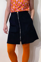 Wearing a colorful patterned top with circular designs, bright orange tights, and the Kubik corduroy zip skirt by BUTIK KUBIK featuring a front zipper, a person confidently stands with one hand in their skirt pocket against a plain white background.