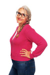 A woman with glasses and long braided gray hair, wearing the BUTIK KUBIK Cissi och Selma cardigan rose and jeans, smiles with one hand on her hip against a white background.