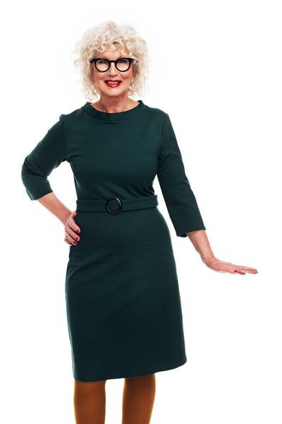 Agneta wears the Cissi och Selma dress Agneta Mörkgrön by BUTIK KUBIK—a dark green, soft viscose dress—paired with brown tights, striking a confident pose with one hand on her hip against a white background.