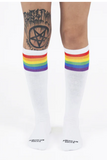 Person bär American Socks Knee High Pride från BUTIK KUBIK – vita, ekovänliga knästrumpor med regnbågsfärgade ränder upptill. Deras knän är synliga, med en stor svart tatuering på det ena knäet. Strumporna har "AMERICAN SOCKS" skrivet vid tårna.