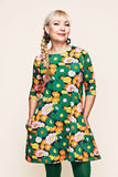 A woman with blonde hair in a side braid wears the BUTIK KUBIK Cissi och Selma Anita 1970 Grönorange, an oversized tunic with pockets featuring a colorful floral design in green, orange, yellow, and pink, paired with green earrings and standing against a plain light background.