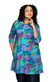 A woman with curly dark hair models the Cissi och Selma Anita Trädgård lila tunic from BUTIK KUBIK, showcasing its vibrant blue, purple, and green floral pattern as she smiles confidently against a white background.