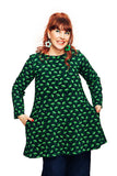 A red-haired woman smiles in the Cissi och Selma Annelie tunic dress, Paraply Grön by BUTIK KUBIK. The ekologisk bomullstrikå dress features light green mushroom patterns, has pockets where she rests her hands, and she wears flower-shaped earrings against a plain white background.