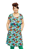 A smiling woman with brown hair in an updo wears the Cissi och Selma dress Asta Humlesurr blå from BUTIK KUBIK, paired with green tights and earrings, standing with her hands in her pockets against a white background.