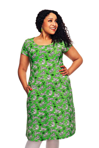 A woman with curly hair and green earrings smiles in the Cissi och Selma dress Asta Tigrar grön from BUTIK KUBIK, a short-sleeved, knee-length green zebra print dress, standing against a white background.