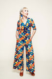 A woman with blonde hair in a braid wears the Cissi och Selma Beata jumpsuit 1970 Blåorange by BUTIK KUBIK, made from stretch ekologisk bomullstrikå and featuring vibrant blue, orange, and yellow floral patterns. She accessorizes with blue earrings and brown shoes, stands with her hands in her pockets, and looks slightly to the side.