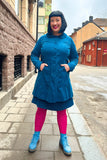 A woman with black hair smiles on a city sidewalk, wearing the BUTIK KUBIK Cissi och Selma coat Berit Petrol and matching skirt in an A-line cut, paired with blue boots and bright pink tights against red and brown buildings.