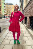 A woman with a blond braid smiles on a city sidewalk, hands in the pockets of the Cissi och Selma coat Berit Ros by BUTIK KUBIK. She wears pink tights, green shoes, and stands before colorful buildings. #kappa60-tal.