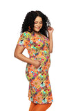 A woman with curly hair smiles and looks down, wearing the Cissi och Selma klänning Betty Fågeltivoli orange by BUTIK KUBIK in a colorful floral pattern, her hand in the pocket as she stands against a white background.
