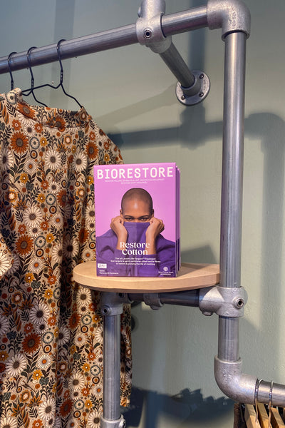 Packets of BUTIK KUBIK Biorestore Restore Cotton laundry detergent are stacked on a round wooden shelf attached to a metal pipe rack, beside a floral-patterned garment hanging on the left.