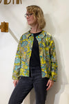A person wearing glasses, a black shirt, dark jeans, and the BUTIK KUBIK Kubik blouse vintage fabric with a colorful floral pattern stands against a white wall, looking to the side.