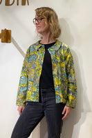 A person wearing glasses, a black shirt, dark jeans, and the BUTIK KUBIK Kubik blouse vintage fabric with a colorful floral pattern stands against a white wall, looking to the side.