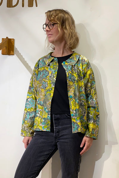 A person wearing glasses, a black shirt, dark jeans, and the BUTIK KUBIK Kubik blouse vintage fabric with a colorful floral pattern stands against a white wall, looking to the side.