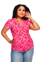 Someone with long curly hair is confidently posed against a white background, wearing the Cissi och Selma top Jessica Nypon Rosa by BUTIK KUBIK. This pink wrap top features a red leaf pattern crafted from LENZING™ ECOVERO™ viscose, paired with blue jeans.