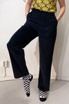 Against a white wall, someone models black Kubik corduroy trousers by BUTIK KUBIK with a colorful shirt. Their raised left foot reveals black-and-white checkered socks. A white electrical outlet is also in view.