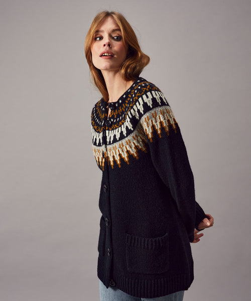 A woman with light brown hair wears the Jumperfabriken Cardigan Cilla from BUTIK KUBIK, made from 100% wool, over light blue jeans. She stands against a gray background, gazing upward with a relaxed expression.