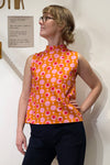 A person wearing glasses models the BUTIK KUBIK Kubik top cineast Popsicle, a sleeveless orange top with a 60s inspired pink and white circular pattern. Made from GOTS certified fabric, they pose confidently with one hand on their hip against a white wall featuring a small Swedish sign.