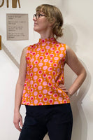 A person wearing glasses models the BUTIK KUBIK Kubik top cineast Popsicle, a sleeveless orange top with a 60s inspired pink and white circular pattern. Made from GOTS certified fabric, they pose confidently with one hand on their hip against a white wall featuring a small Swedish sign.