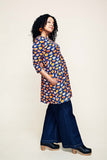 Wearing the BUTIK KUBIK Cissi och Selma Anita tunic “Kittens koppar orange,” a woman stands sideways with her hands in her pockets, smiling confidently in wide-leg jeans and clogs.