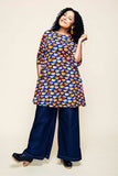 A woman with curly hair smiles, wearing the BUTIK KUBIK Cissi och Selma Anita tunic Kittens koppar orange—with its colorful pattern and practical pockets—paired with wide-legged dark jeans and black clogs, standing against a plain background.