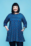 A woman with short black hair smiles, standing against a blue background. She wears the Cissi och Selma Anita tunic Rävar by BUTIK KUBIK—an oversized blue tunic with a red pattern—and has her hands in her pockets. Her right forearm features a colorful tattoo.