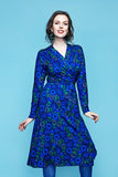 A woman wears a Cissi och Selma Anna-Klara dress Hassel Blå, a long-sleeved, blue omlottklänning in LENZING ECOVERO viscose jersey with a repeating pattern of green and blue stylized flowers and leaves.