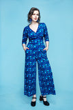 A woman wearing a blue Cissi och Selma Beata jumpsuit Droppe blå in a wrap-style with a repeating circular pattern and three-quarter length sleeves, standing with her hands in her pockets.