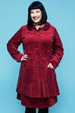 A person with black hair and bangs smiles while wearing the Cissi och Selma coat Berit Manchester rabarber from BUTIK KUBIK, featuring large buttons and patch pockets, against a light blue background.