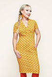 A woman with a side braid wears the Cissi och Selma klänning Betty Miniblom senap by BUTIK KUBIK, a figure-hugging summer dress in a yellow patterned fabric, paired with orange flower earrings and red tights, standing against a plain light background and smiling slightly.