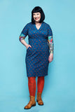 Woman with black hair wearing the Cissi och Selma klänning Betty Rävar from BUTIK KUBIK—a fitted summer dress in a blue pattern—paired with orange tights, brown boots, and displaying colorful arm tattoos, stands against a light blue background and smiles at the camera.