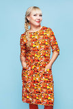 A smiling woman with blond hair in a side braid wears the Cissi och Selma dress Doris Flugsvamp by BUTIK KUBIK, featuring orange, yellow, and white 60s-inspired patterns in eco-friendly cotton jersey. She pairs the dress with matching orange earrings and stands against a light blue background with her hands in her pockets.