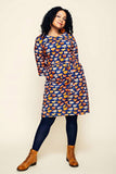 Woman with curly hair wearing the Cissi och Selma dress Doris Kittens koppar orange from BUTIK KUBIK, paired with black tights and brown lace-up boots, standing against a beige background.