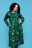 A woman with curly dark hair wears a Cissi och Selma dress Helga Kaprisblomma grön, a long-sleeved green midi dress in soft viscose jersey featuring a pattern of small white caper flowers and vines.