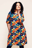 A person with curly hair smiles confidently, wearing the Ingrid 1970 Blåorange dress from BUTIK KUBIK’s Cissi och Selma collection—a colorful, floral-patterned piece adorned with red, orange, yellow, and blue flowers. With one hand in their pocket, they highlight the practical design of this klänning med fickor as they stand against a plain light background.