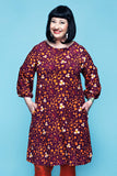 A smiling woman wearing a Cissi och Selma dress Ingrid Kaprisblomma plommon, a knee-length, plum-colored organic cotton dress with a repeating orange and white floral print and three-quarter length sleeves.