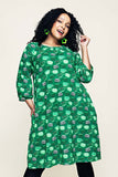 A smiling woman with curly hair wears the Cissi och Selma dress Ingrid Kittens koppar grön from BUTIK KUBIK, featuring a green patterned stretch design with three-quarter sleeves and paired with green earrings, standing against a plain background.