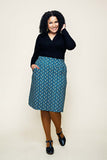 A woman with curly hair smiles, wearing a black long-sleeve top, the Cissi och Selma Majken skirt Miniblom blåbär from BUTIK KUBIK—a patterned blue pennkjol i ekologisk bomull with praktiska fickor—brown tights, and black shoes. She stands against a plain background with one hand in her pocket and the other touching her hair.