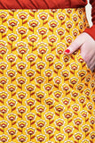A person wearing the Cissi och Selma Majken skirt Miniblom senap from BUTIK KUBIK—with its yellow, white, and red retro floral pattern—rests their right hand in one of the pockets. Their shirt and nail polish are both red, and the skirt is crafted from ekologisk bomull for a sustainable touch.