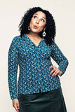 A woman with curly hair wears the Cissi och Selma blus Matilda Miniblom blåbär from BUTIK KUBIK—a blue patterned long-sleeve blouse—and a green skirt. She stands with one hand on her hip, looking up with a playful expression. She has white earrings, red nail polish, and is posed against a plain light background.