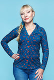 A woman with blonde braided hair, wearing the Cissi och Selma blus Matilda Rävar from BUTIK KUBIK—a blå dam skjorta with a red pattern—paired with blue earrings and jeans, stands against a light blue background, smiling at the camera.
