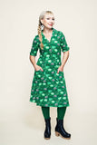 A woman with a long blonde braid wears a BUTIK KUBIK Cissi och Selma dress Monica Kittens koppar grön, paired with green tights and black heeled shoes, standing with her hands in her dress pockets against a plain light background.