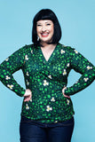 A woman with short dark hair smiling, wearing a green, v-necked wrap blouse in the Cissi och Selma blus Susanne Kaprisblomma grön style, featuring a white flower and vine pattern.