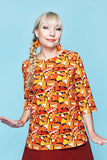 Smiling person with blond braided hair wears the Cissi och Selma top Vendela Flugsvamp by BUTIK KUBIK—an orange ekologisk bomullstopp with a mushroom pattern—paired with orange earrings and a red skirt, standing against a light blue background.