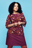A person wearing a Cissi och Selma top Vendela Kaprisblomma plommon in organic cotton with an orange and white floral pattern and a matching plum-colored A-line skirt.