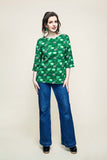 A woman stands against an off-white background wearing the “Cissi och Selma top Vendela Kittens koppar grön” by BUTIK KUBIK, styled with blue flared jeans, dark clogs, and earrings. Her dark hair is worn down and she poses with one hand on her hip.