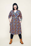 A smiling person with black hair wearing the Cissi och Selma dress Vera Kittens koppar orange from BUTIK KUBIK, featuring a patterned design and three-quarter sleeves, stands against a plain light background with hands in the dress pockets. They pair the dress with black leggings and brown boots.